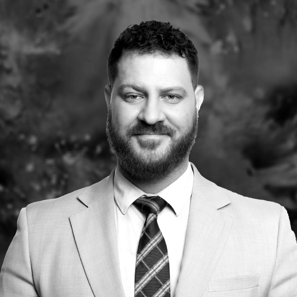 John Paul Fraser, a man with a beard and curly hair, smiles in a light suit and plaid tie in a professional headshot.