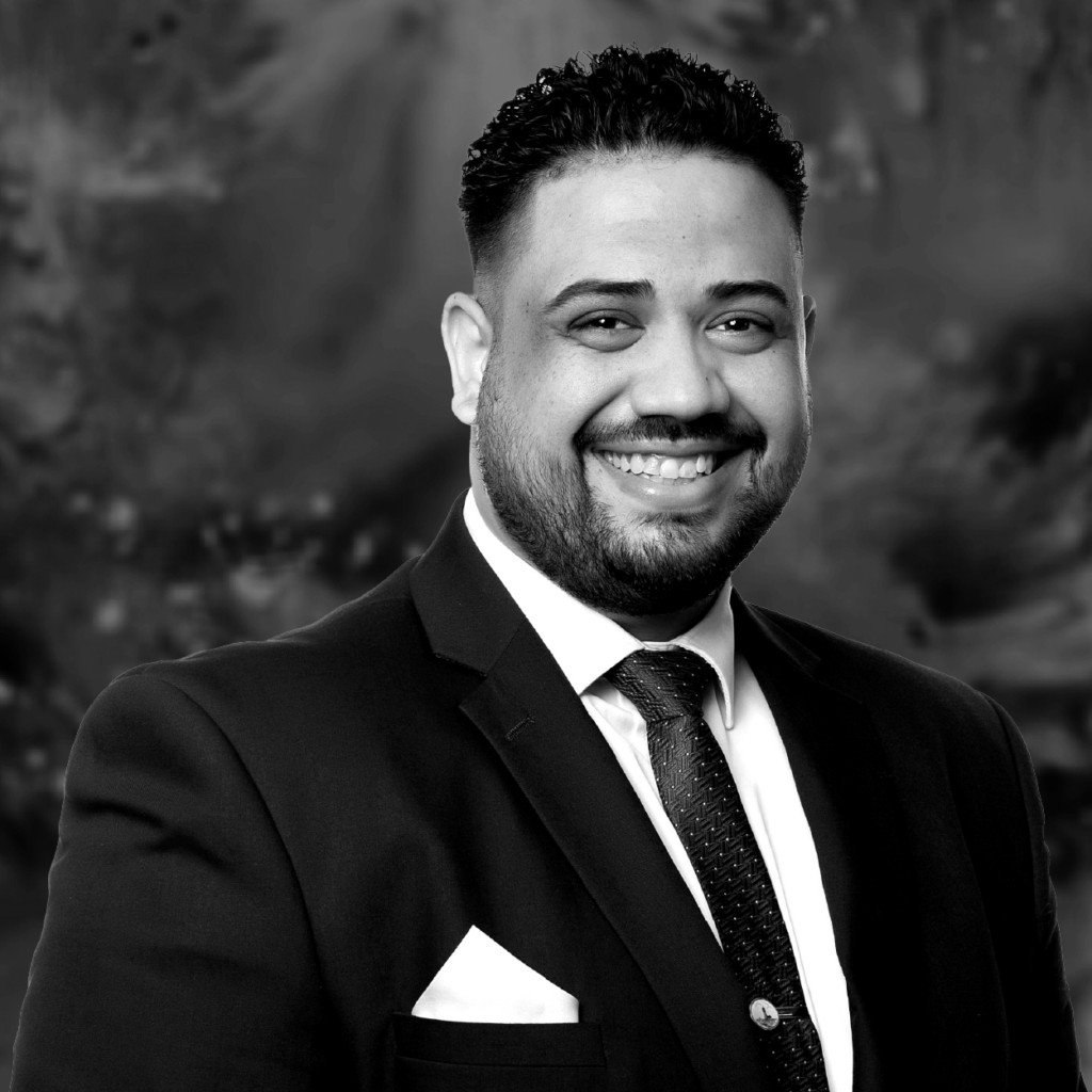 Professional headshot of a smiling man with a beard and short curly hair, wearing a suit and tie.